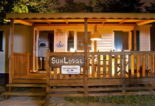 Avondfoto van een verlichte houten lodge met veranda, SunLodge-bord en gezellige sfeer.