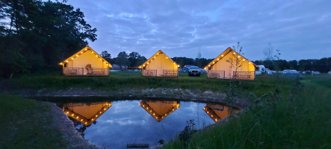 Lit safari tents at Beloofde Land, Netherlands, glowing by a pond at dusk, reflected in tranquil water.
