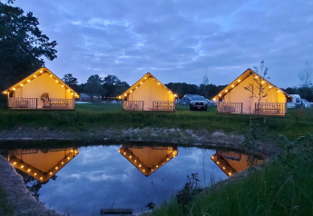 Lit safari tents at Beloofde Land, Netherlands, glowing by a pond at dusk, reflected in tranquil water.