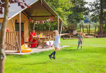 Children play badminton in front of a safari tent at Beloofde Land, Netherlands, with woman watching.