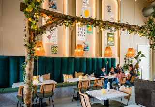 Een modern glampingrestaurant met groene banken, houten decoratie en een gezin dat samen aan tafel zit.