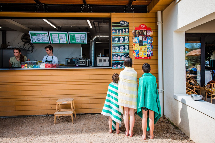 Tres niños con toallas eligen helados en un quiosco de una tienda de safari en un campamento de verano.