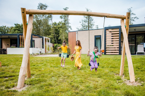 Drei Kinder spielen und schaukeln draußen vor einem Safarizelt mit viel Grün und modernen Gebäuden.
