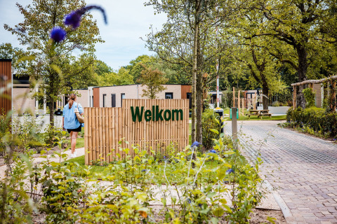 Entrance to a safari tent resort with a welcome sign, greenery, and a person walking towards the lodges.