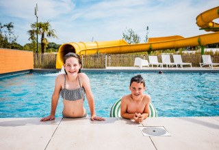 Two children enjoy a swimming pool at a glamping site, with a yellow water slide and lounge chairs behind them.