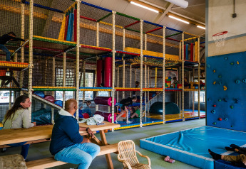 Parque infantil cubierto con estructura de escalada, pared de rocas y adultos sentados en mesas en Beloofde Land.