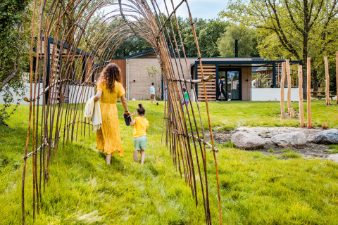 A woman and child walk through a natural archway on a grassy path towards a modern glamping accommodation.