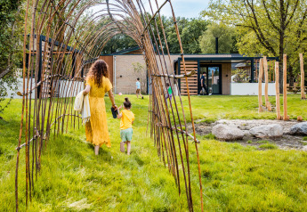 Mujer y niña caminan por un túnel natural de ramas hacia un alojamiento de glamping moderno al aire libre.