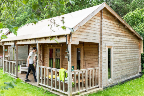 Photo of a wooden safari tent cabin with porch in a green setting, people relaxing on the terrace outside.