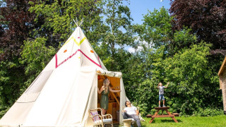 Gezin ontspant voor de Jippi Tippi tipi tent, omringd door groen gras en hoge bomen in de natuur.