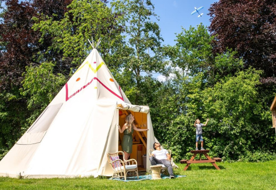 Gezin geniet buiten bij de Jippi Tippi tipi-tent, omringd door bomen en gras op een zonnige dag.
