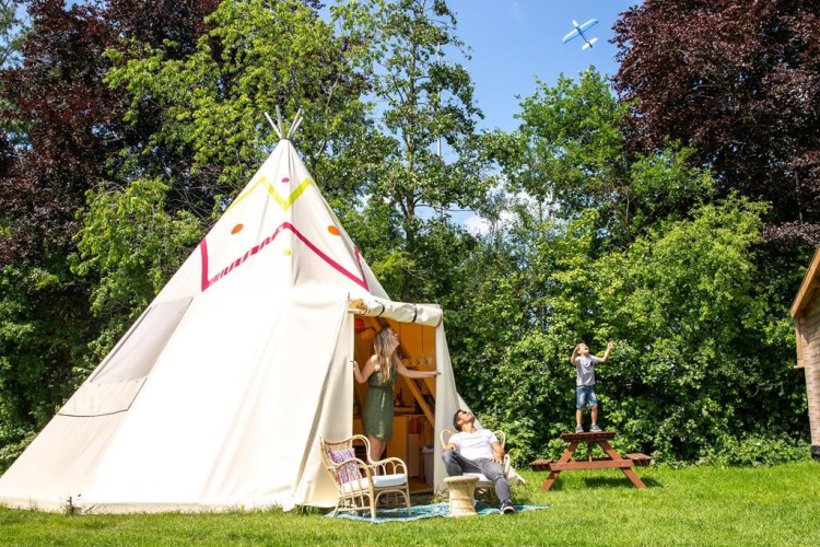 Gezin geniet buiten bij de Jippi Tippi tipi-tent, omringd door bomen en gras op een zonnige dag.