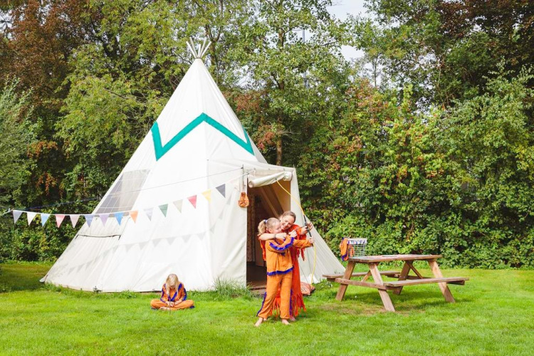 Kinderen in oranje kostuums spelen bij grote tipi en picknickbank op Beloofde Land, Nederland.