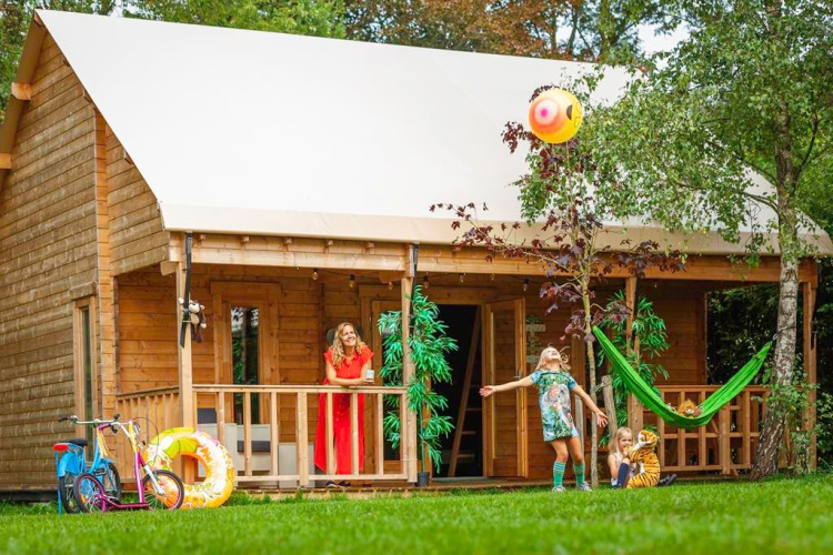 Familie entspannt vor einem Jungle huisje Safari-Zelt, während Kinder auf dem Rasen mit einem Ball spielen.
