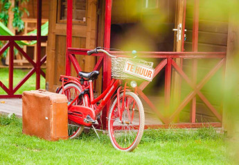 Bicicletta rossa con cartello 'TE HUUR' e valigia marrone fuori da Zweedse Huisjes al Beloofde Land, Olanda.