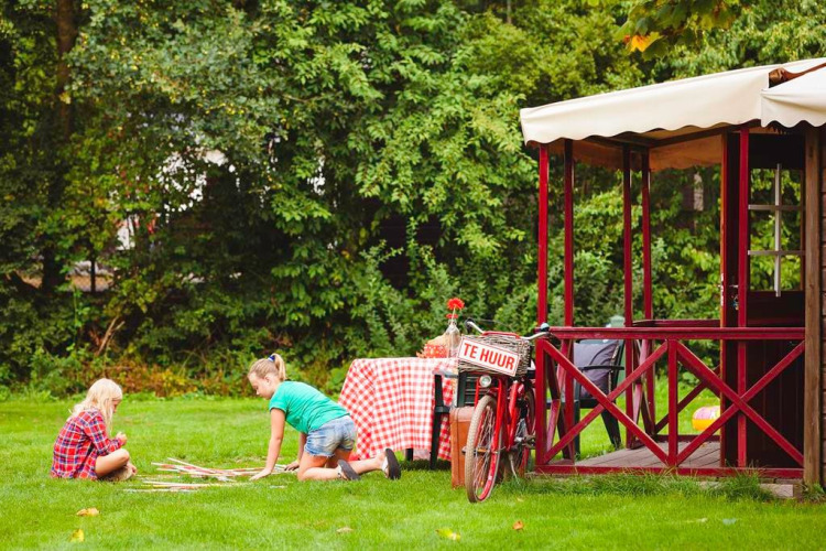 Zwei Kinder spielen auf dem Rasen neben einer Hütte und einem Fahrrad bei Zweedse Huisjes, Beloofde Land.