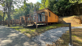 Houten lodge met terras in een bosrijke omgeving, zonnige dag, verlicht grindpad en zitplaatsen buiten.