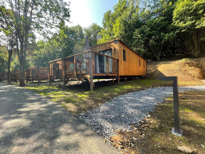 Houten lodge met terras in een bosrijke omgeving, zonnige dag, verlicht grindpad en zitplaatsen buiten.
