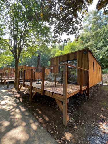 Wooden ecolodge at Hameau de l'Ourthe, Belgium, with deck seating and surrounded by lush greenery.
