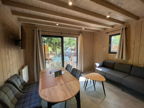 Inde i en hyggelig lodge med træpaneler, sofa, spisebord og udsigt til en terrasse gennem store vinduer.