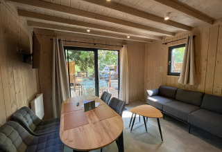 Inde i en hyggelig lodge med træpaneler, sofa, spisebord og udsigt til en terrasse gennem store vinduer.