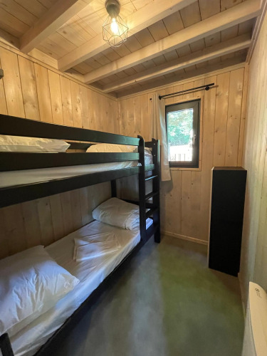 Pequeño dormitorio en una cabaña con paredes de madera, litera, ventana, cortina y lámpara de techo.