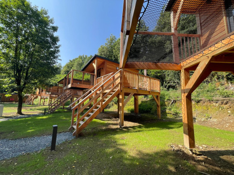 Bomenhutten op palen met touwbrug bij Hameau de l'Ourthe, België, omgeven door bomen en grasveld.