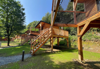 Casas en los árboles sobre pilotes con puente de red en Hameau de l'Ourthe, Bélgica, rodeadas de verde.