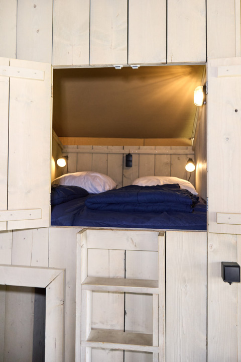 Ingresso a una zona notte accogliente in una tenda safari con pareti in legno, scala e lenzuola blu.