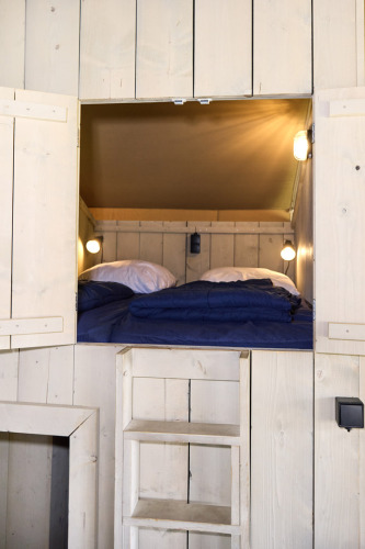Entrada a una acogedora zona de dormir en tienda safari con paredes de madera, escalera y ropa azul.