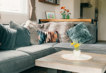Stilfuld lounge i Veluwe-lodge med hjørnesofa, puder og blomster på bord i De Boshoek feriepark, Holland.