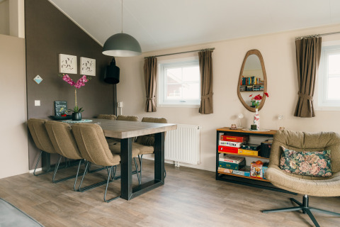 Moderne lodge-stue med spisebord, seks stole, lænestol, bogreol og vinduer på feriepark De Boshoek, Holland.