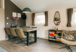 Moderne lodge-stue med spisebord, seks stole, lænestol, bogreol og vinduer på feriepark De Boshoek, Holland.