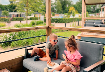 Due bambini mangiano sulla terrazza di una Tiny Lodge al Holiday park De Boshoek, Paesi Bassi, immersi nel verde.