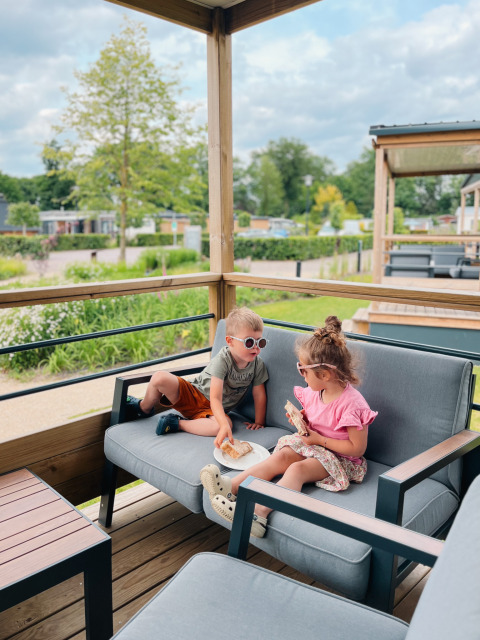 Due bambini mangiano sulla terrazza di una Tiny Lodge al Holiday park De Boshoek, Paesi Bassi, immersi nel verde.