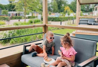 Dos niños comen en la terraza de la Tiny Lodge en Holiday park De Boshoek, Países Bajos, rodeados de verde.
