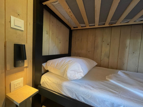 En enkel seng med træpaneler, hvidt sengetøj, hylde, stikkontakt og lampe på et lodgeværelse.