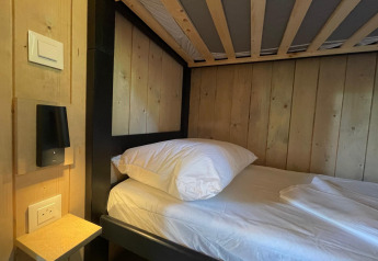 Een eenvoudig eenpersoonsbed met houten panelen, wit beddengoed, plankje, stopcontact en lamp in een lodge.