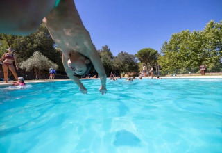Child jumps into pool at Camping Begur, a holiday park in Catalonia, Spain, surrounded by trees and people.