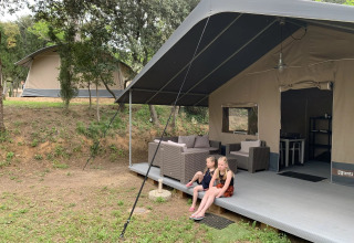 Twee kinderen zitten voor een safaritent op Camping Begur in Catalonië, Spanje, tussen de bomen.