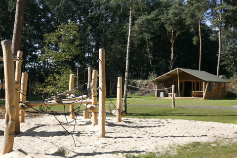 Spielplatz mit Klettergerüst aus Holz und Sand vor einer Safarilodge Deluxe im grünen Waldgebiet.