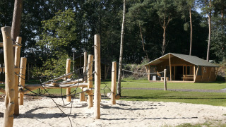 Parco giochi con struttura in legno e sabbia vicino a un’elegante tenda safari in una zona boschiva.
