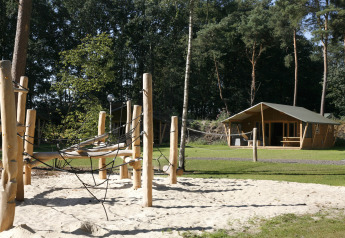 Aire de jeux avec structure bois et sable devant une tente safari lodge de luxe, en forêt verdoyante.