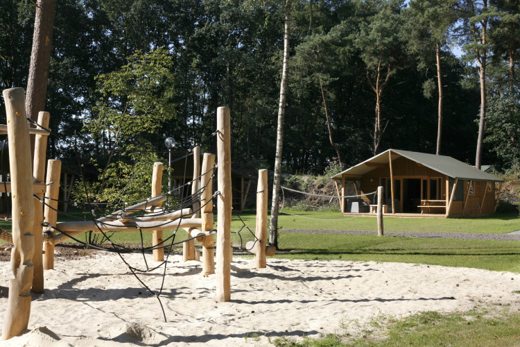 Aire de jeux avec structure bois et sable devant une tente safari lodge de luxe, en forêt verdoyante.