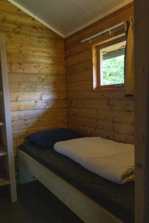 Gemütliches Holz-Zimmer mit Einzelbett, Fenster und Vorhang in einer Safarilodge Deluxe in den Niederlanden.