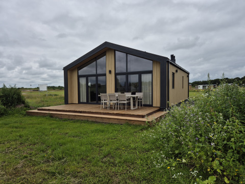 Lodge Type Dommel avec grandes fenêtres et terrasse en bois, entourée de verdure sous un ciel nuageux.