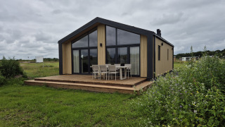 Type Dommel-lodge met ruime ramen en houten terras tussen groene weiden onder een bewolkte hemel.