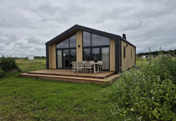 Type Dommel lodge with large windows and outdoor dining set on a wooden deck, surrounded by green fields.