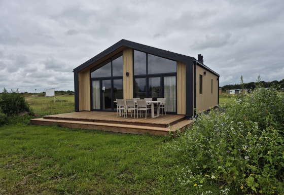 Lodge Type Dommel avec grandes fenêtres et terrasse en bois, entourée de verdure sous un ciel nuageux.