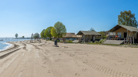 Safari tents at Zuiderzee beach, Netherlands, with trees, playground, clear blue sky, and calm water.
